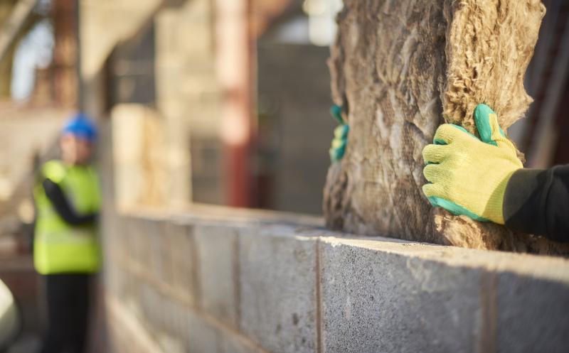 Close up of a worker's gloved hands installing cavity insulation in a wall, colleague in the background