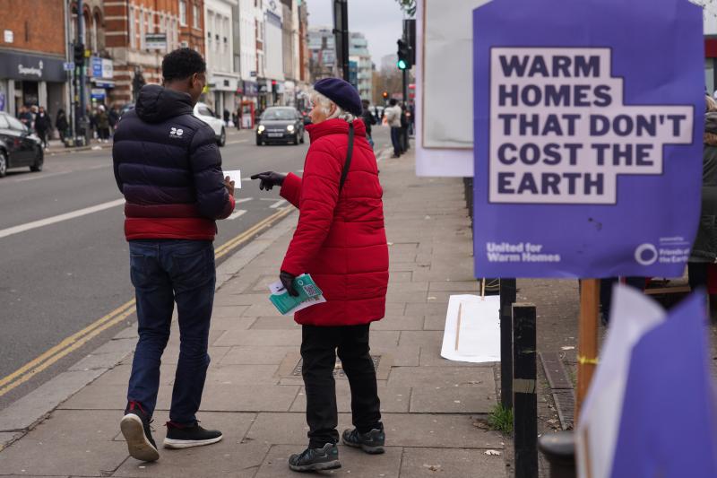 A warm homes campaigner hands a leaflet to a passer-by. In the foreground a placard reads "Warm homes that don't cost the earth"