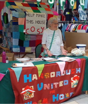 Campaigner stands at a stall with a giant knitted house behind them and a sign that says "Help make this house cosy"