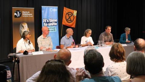 A panel of candidates at a table, with people watching in the foreground and group banners in the background