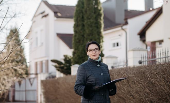 Woman wearing coat and gloves standing outside some homes holding a clipboard