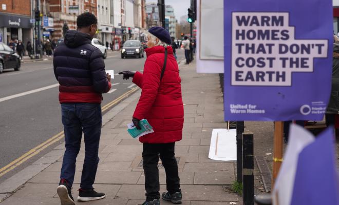 A warm homes campaigner hands a leaflet to a passer-by. In the foreground a placard reads "Warm homes that don't cost the earth"