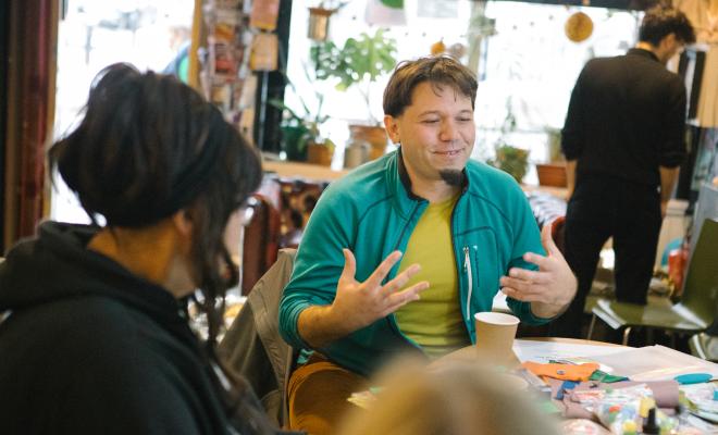 A man is talking, smiling and gesturing at a table in a cafe with a couple of other people visible